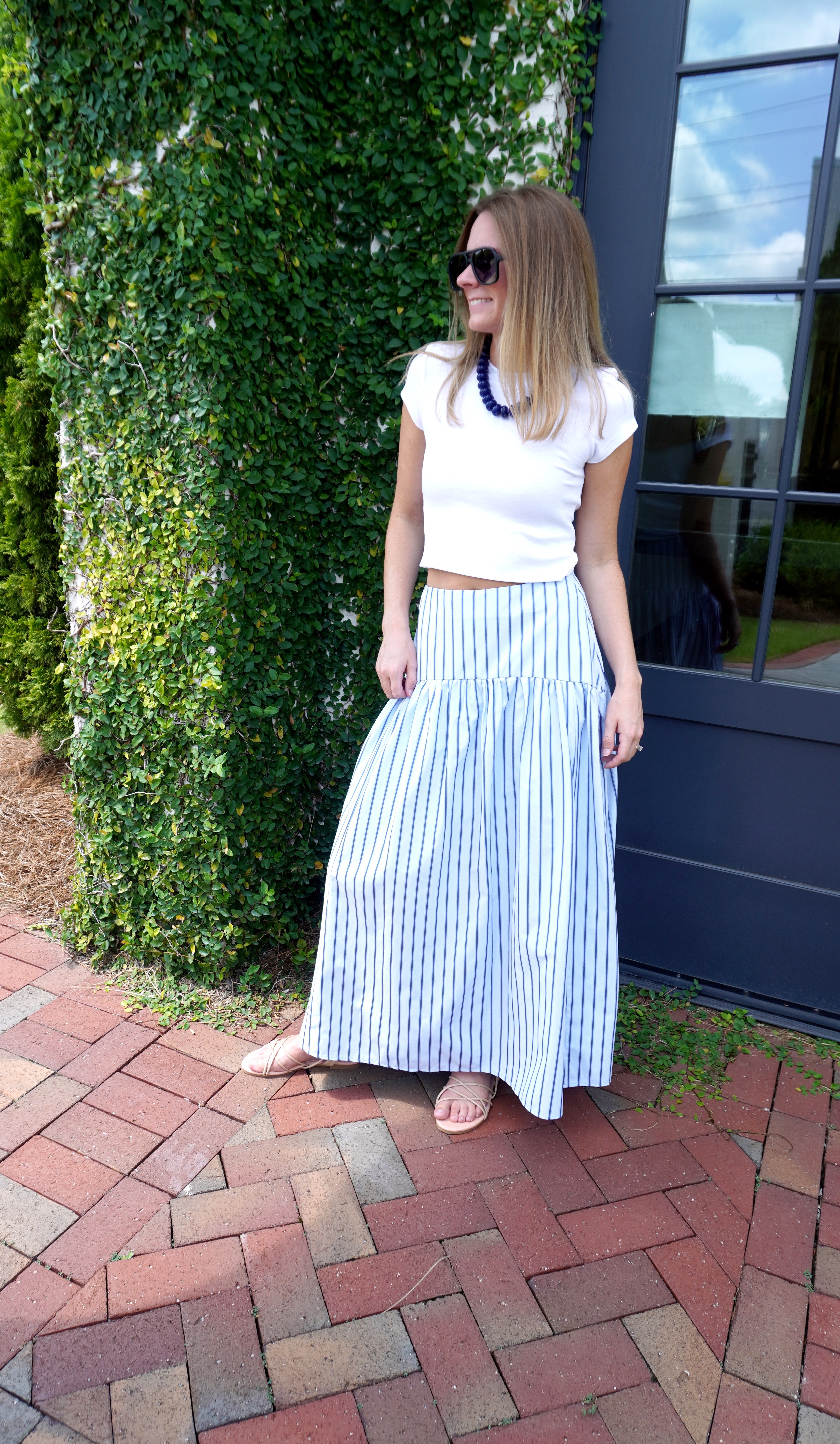 Woman wearing a white fitted short-sleeve top with a blue and white striped maxi skirt, navy statement necklace, and black sunglasses