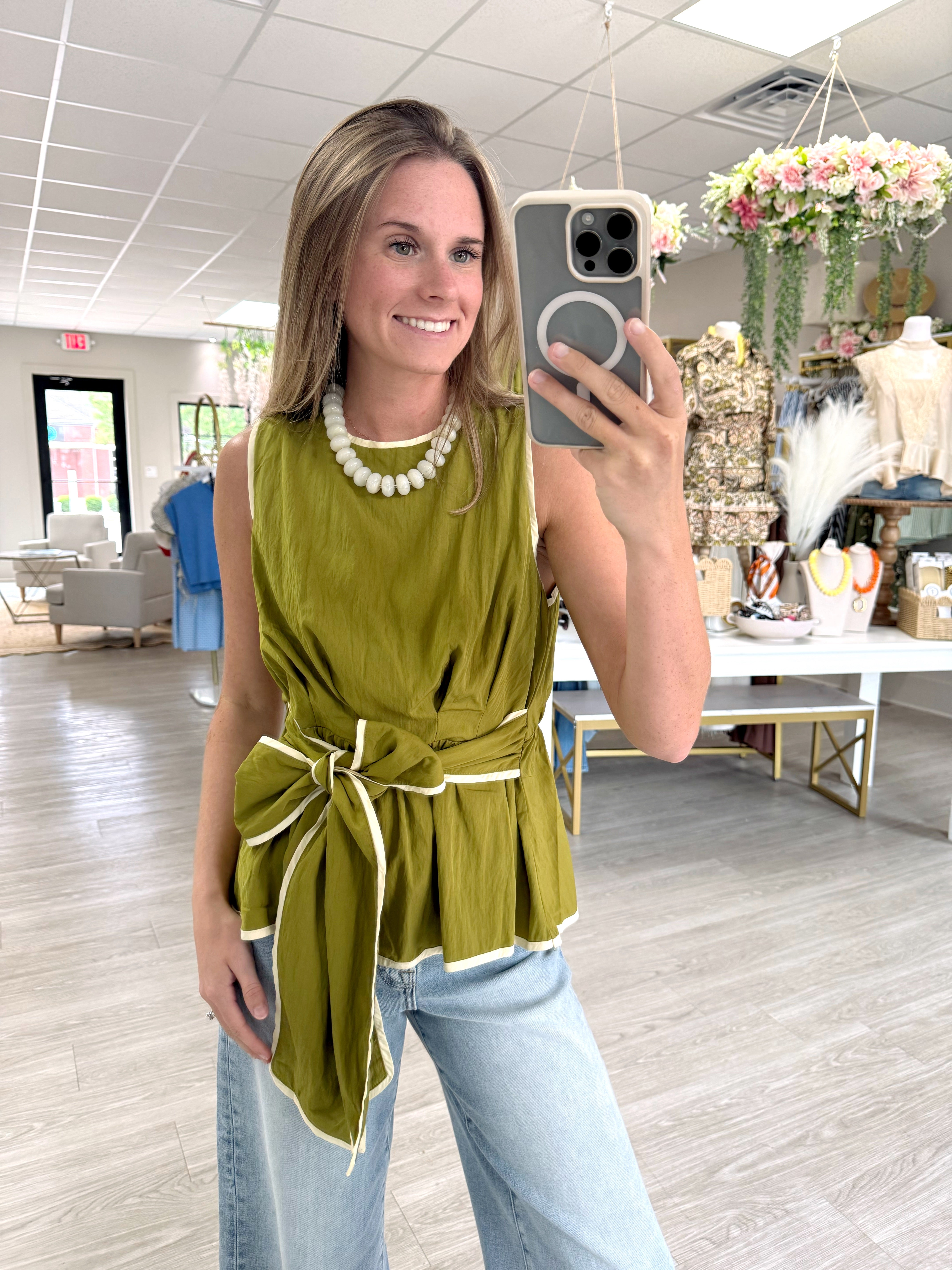 Woman wearing an olive green sleeveless tie waist top with cream contrast trim, styled with light wash wide leg jeans.