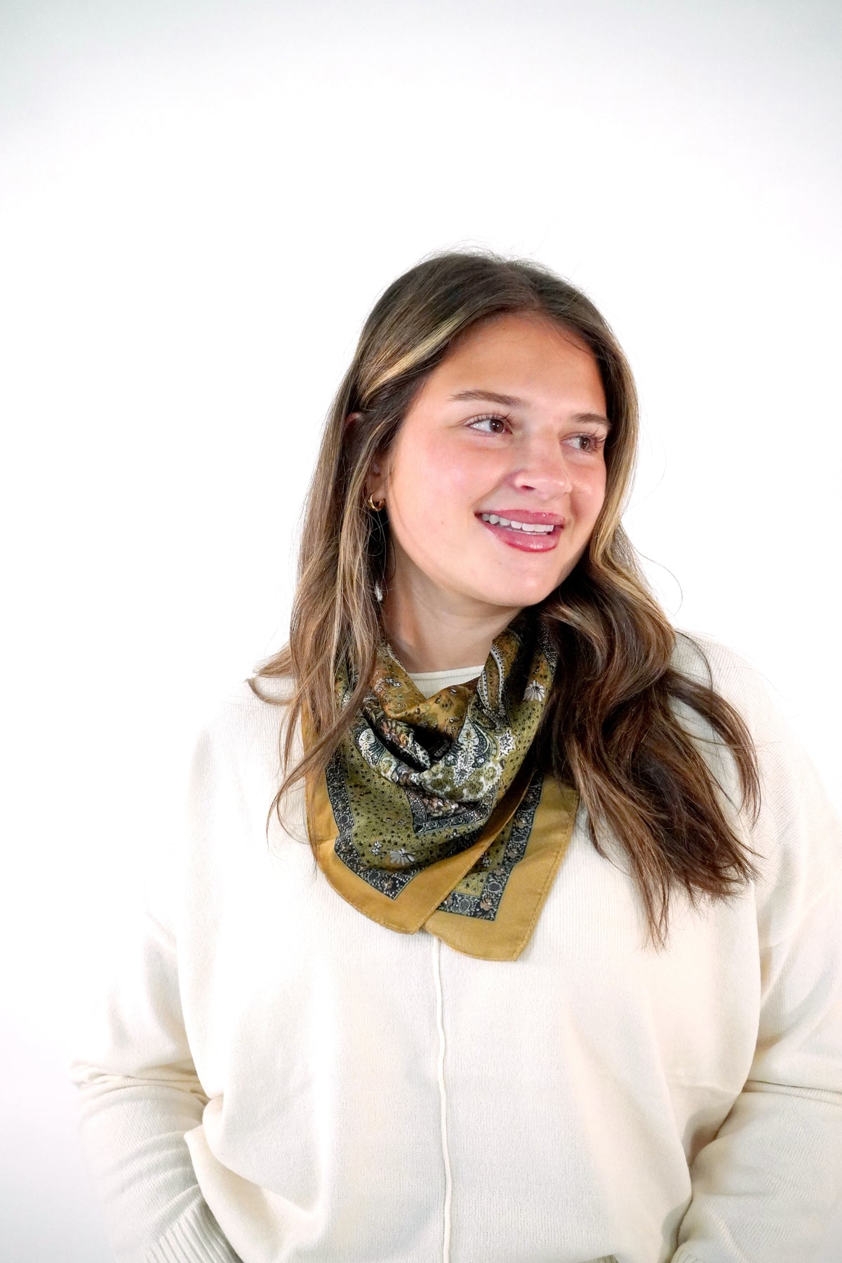 Olive green and camel paisley print satin square scarf tied at the neck over a cream sweater