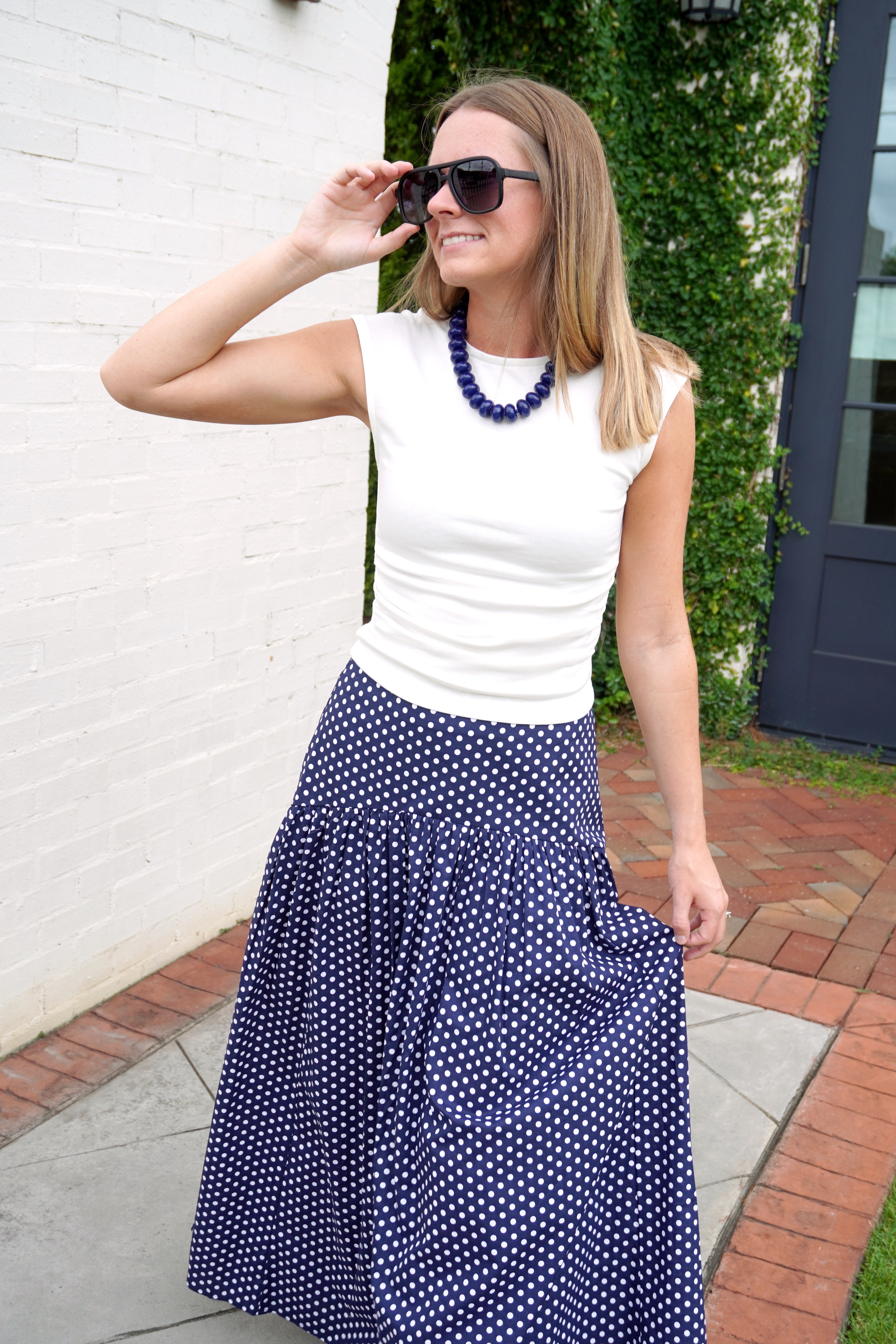 Model wearing a fitted white cap sleeve top with a clean crew neckline, paired with a navy polka dot skirt, navy beaded necklace, and oversized black sunglasses. The classic neutral top offers a sleek, versatile look perfect for everyday styling or layering.