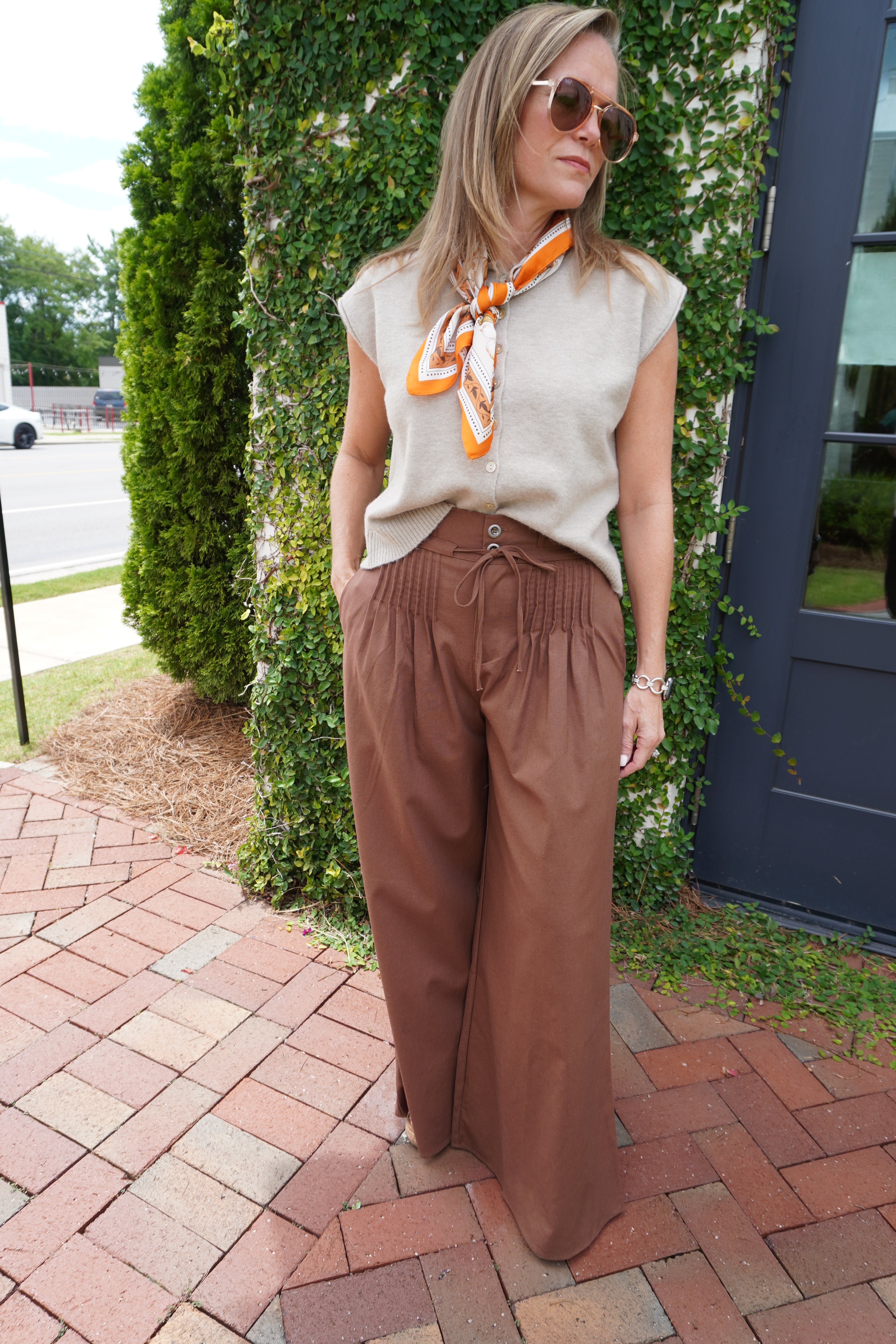 Woman wearing a sleeveless oatmeal button-front knit sweater top styled with brown wide-leg pants and an orange printed neck scarf