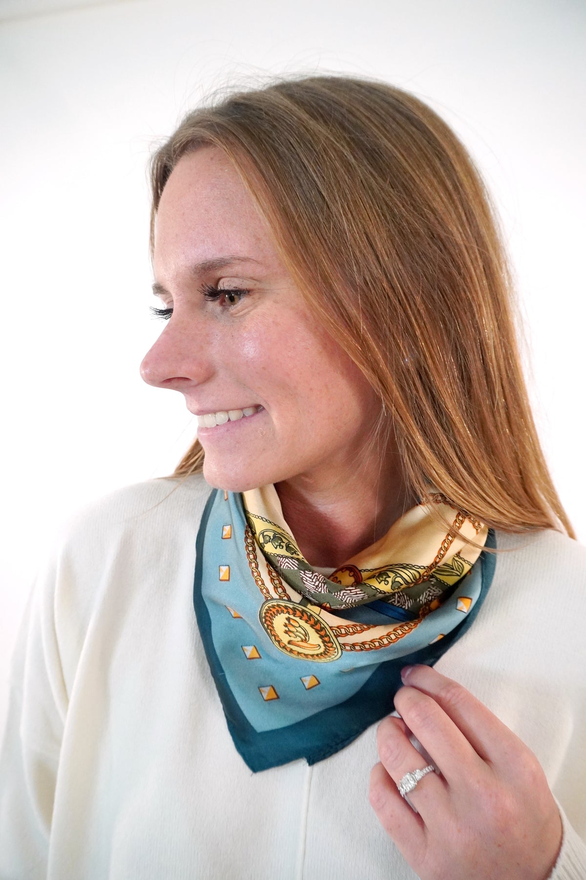 Teal and blush chain print satin square scarf tied at the neck over a cream sweater