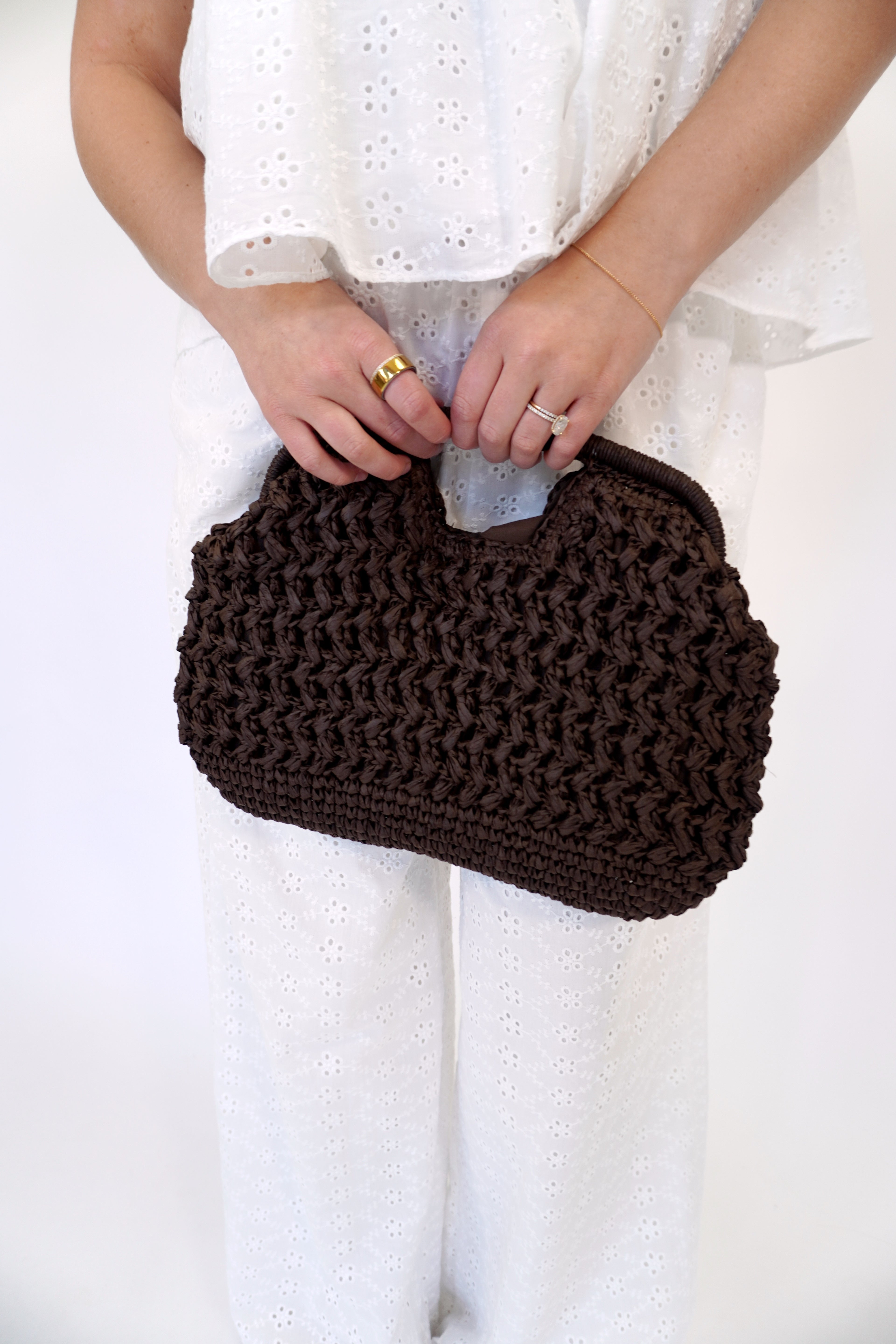 Dark brown woven straw handbag with chunky braided texture and rounded top handle, styled with a white eyelet set and gold jewelry