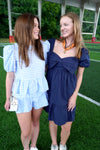 Two women standing on a grassy field, one in a blue checkered dress and the other in a navy dress with a bow, smiling.