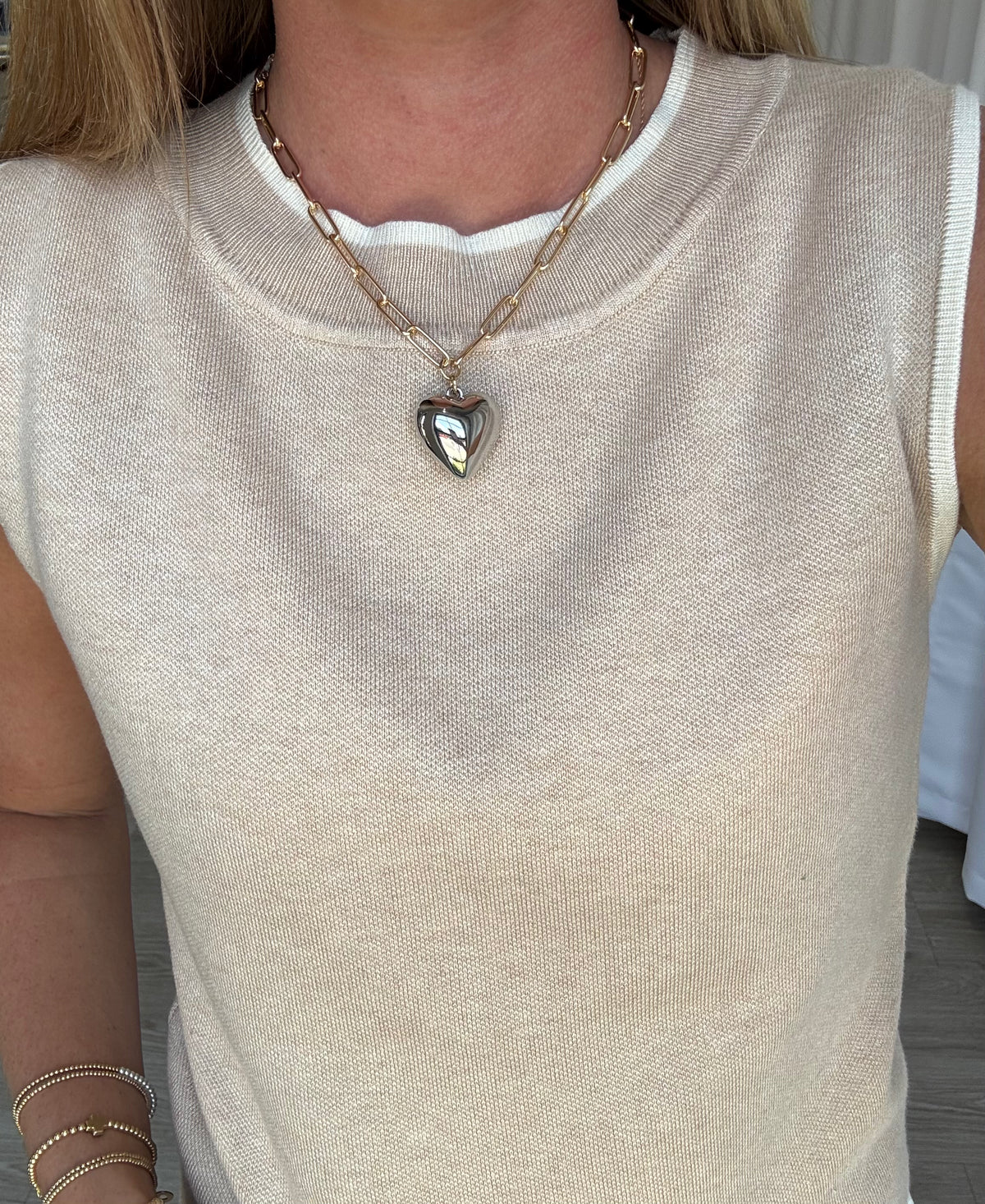 Person wearing a beige sleeveless top with a heart-shaped pendant necklace.