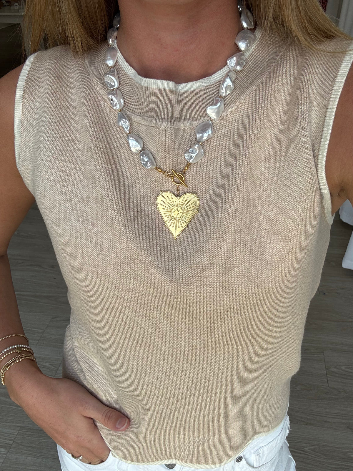 Person wearing a beige sleeveless top with a pearl necklace and gold heart pendant.