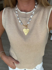 Person wearing a beige sleeveless top with a pearl necklace and gold heart pendant.