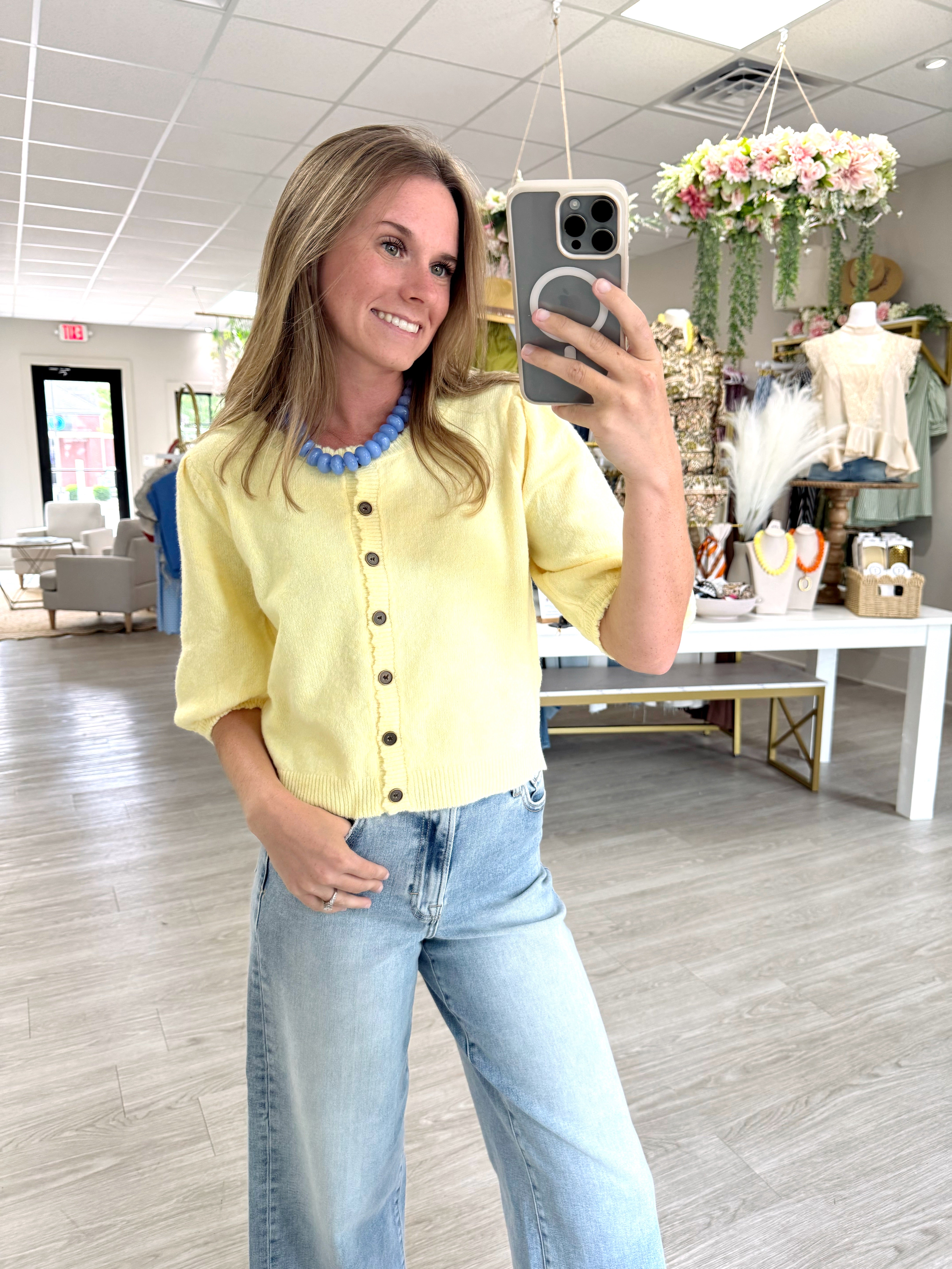 Woman wearing a butter yellow short sleeve button front cardigan sweater styled with light wash jeans and a blue statement necklace.